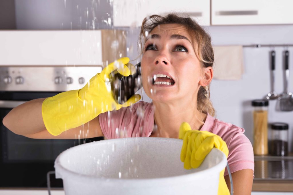 Woman holding a bucket under a leak coming from the ceiling while she’s on the phone with a plumber