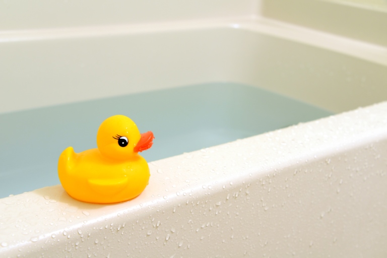 Yellow rubber duck sitting on edge of bathtub with standing water due to a clogged drain.