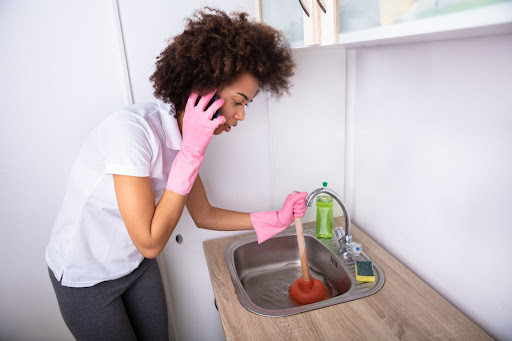 A woman on the phone while she tries to use a plunger in a clogged kitchen sink.