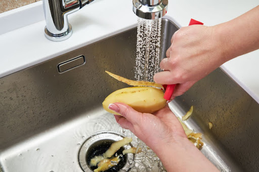 A person peeling a potato over a kitchen sink garbage disposal.